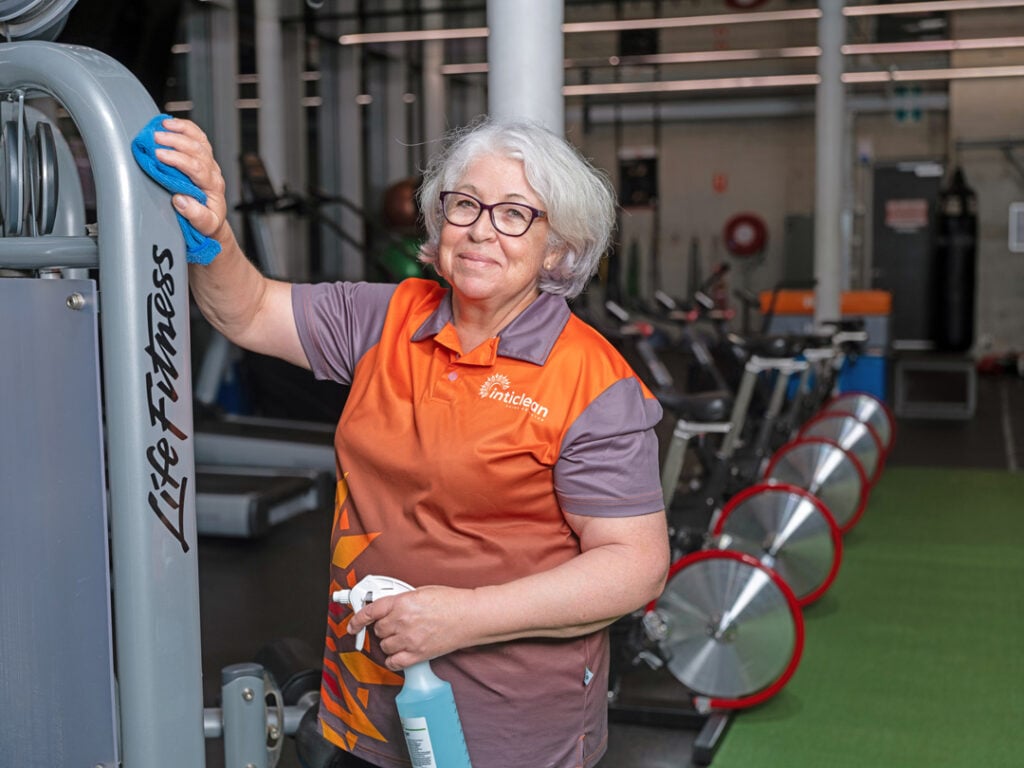 IntiClean staff member cleaning gym equipment with cloth and spray bottle inside fitness centre