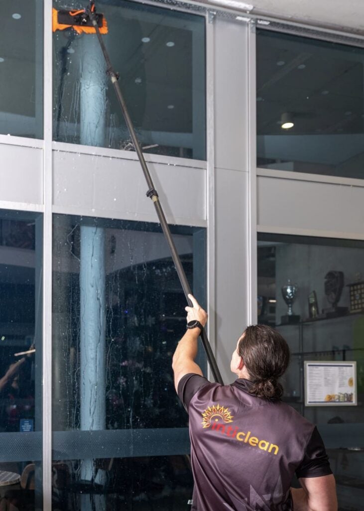 IntiClean worker cleaning tall glass window with extension pole inside modern commercial building