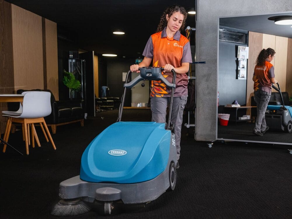 Inticlean cleaning services staff operating Tennant floor sweeper machine on dark carpeted office area