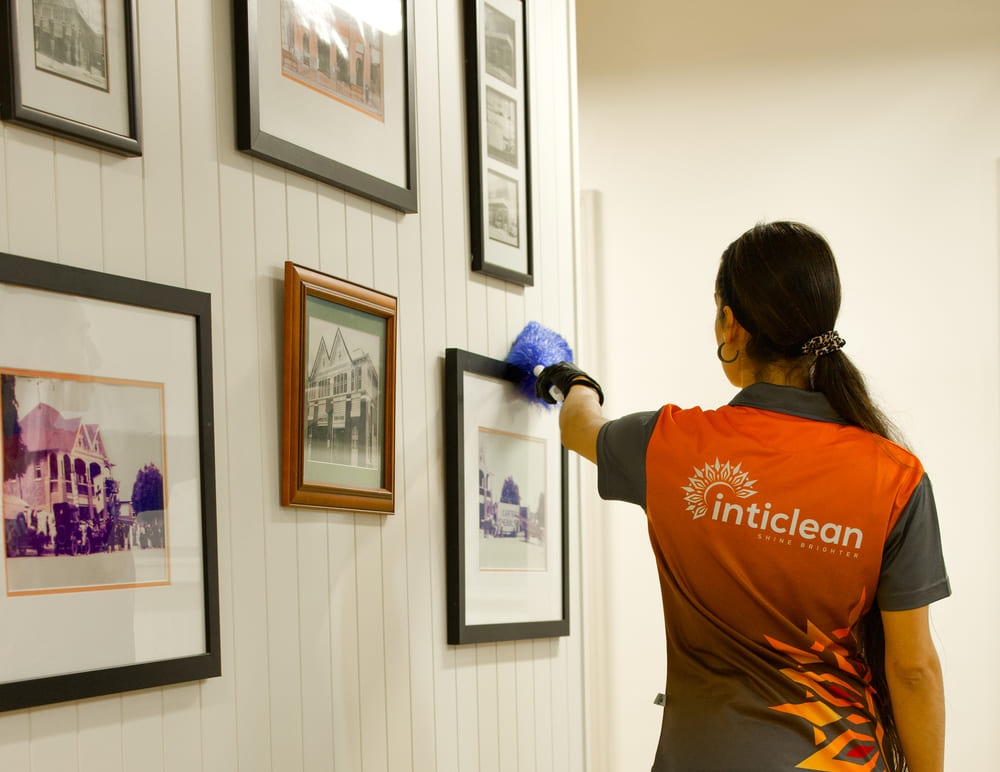IntiClean worker dusting framed photographs on an interior wall with a cleaning tool